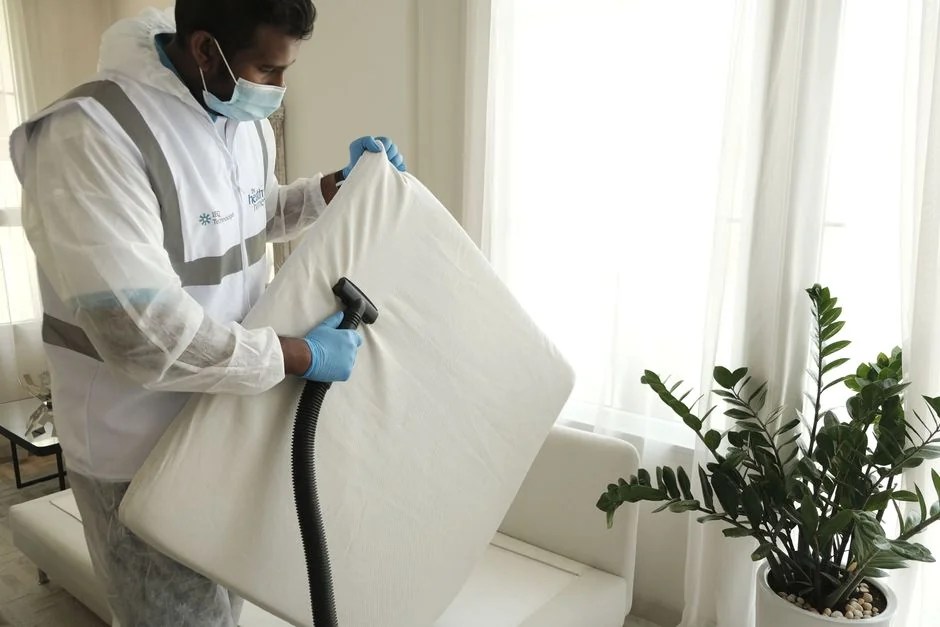 A worker dressed in a safety vest cleans a blue carpet outdoors with a brush.